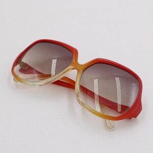 May include: Square-framed sunglasses with a gradient lens. The frames transition from red to yellow, with a clear lower section. The lenses are a gradient brown. A vintage accessory.