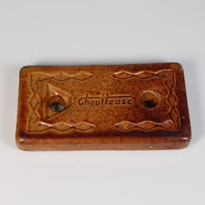 May include: A rectangular, brown ceramic object with the word "Choutteuse" in the center. The object has two holes and decorative patterns around the edges. The ceramic piece is likely a vintage or antique item.
