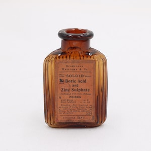 K&ouml;nnte beinhalten: Eine bernsteinfarbene Glasflasche mit braunem Etikett. Auf dem Etikett steht "SOLOID Boric Acid and Zinc Sulphate" und das Wort "POISON". Die Flasche hat ein geripptes Design und einen dunkelbraunen Verschluss.