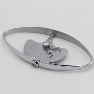 Könnte beinhalten: Ein silberfarbenes Metallarmband mit ovalem Design. Das Armband hat ein dekoratives Element mit kreisförmigem Design und einem kleinen, runden Juwel. Das Armband ist vor einem weißen Hintergrund platziert.