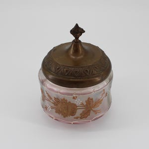May include: An antique jar with a pink tint and a decorative brass lid. The jar has a floral design in gold and white. The lid features an ornate pattern and a small finial. The jar is likely for storing small items, such as trinkets or powders.