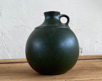 May include: A dark green ceramic vase with a round body and a single handle. The vase is sitting on a wooden surface.