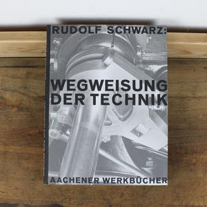 May include: A book titled "Rudolf Schwarz: Wegweisung der Technik" with a black and white image of machinery on the cover. The book is on a wooden surface.