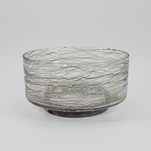 May include: A clear glass bowl with a unique design. The bowl has a cylindrical shape with a slightly flared rim and a dark base. The exterior is decorated with a web-like pattern of thin, gray lines, creating a textured appearance. The bowl is likely used for decorative purposes.