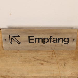 May include: A wooden sign with the word "Empfang" in black letters and an arrow pointing left. The sign is mounted on a light-colored wooden surface, with a white wall in the background.