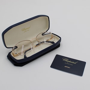 May include: Gold-rimmed eyeglasses in a navy blue case with a cream interior. The case is open, displaying the glasses. A navy blue card with gold text and the brand name "Chopard" is next to the case.