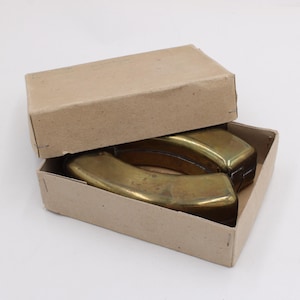 May include: Two brass-colored, curved objects are inside a partially open, rectangular cardboard box. The box is light brown. The objects have a rounded shape and appear to be made of metal. The box is on a white surface.