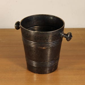 May include: A dark gray metal ice bucket with two small handles. The bucket has horizontal bands around the body and a textured surface. It is sitting on a wooden surface.