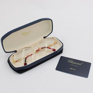 Könnte beinhalten: Goldgerahmte Brille mit roten Details in einem offenen marineblauen Etui. Eine marineblaue Karte mit goldfarbenen Buchstaben, einschließlich der Marke "Chopard", liegt neben dem Etui. Das Innere des Etuis ist cremefarben.