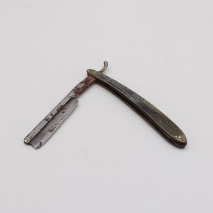 Peut inclure: Un rasoir droit ancien avec un manche incurvé et une lame partiellement ouverte. La lame montre des signes d'âge et d'usure, avec de la rouille visible. Le manche est sombre et semble être en bois ou dans un matériau similaire.