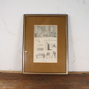 May include: Framed antique print with a gold-colored frame. The print features detailed black and white illustrations of people and objects, likely from a historical or scientific context. The frame is resting on a rustic wooden surface against a white wall.