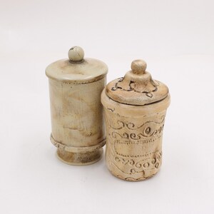 May include: Two beige ceramic jars with lids. The taller jar is cylindrical with a simple lid and a small knob. The shorter jar has a decorative lid and etched patterns around the body. Both jars are on a white background.