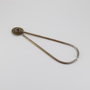 Op de afbeelding: Een vintage metalen gereedschap met een cirkelvormige messing kop en twee gebogen armen. De armen zijn gemaakt van een messingkleurig metaal dat aan de uiteinden overgaat in een zilverkleur. De tool wordt waarschijnlijk gebruikt om te meten of te markeren.