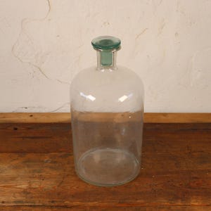 May include: A clear glass bottle with a rounded body and a green glass stopper. The bottle sits on a rustic wooden surface. The background is a textured, off-white wall.