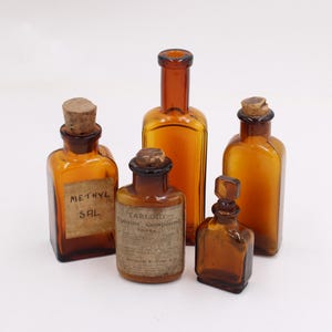 May include: Five antique amber glass bottles with cork stoppers. Two bottles have labels that read "METHYL SAL" and "CARBOLIC POISONING COMPOUND". The bottles vary in shape and size, with a square, rectangular, and cylindrical design.