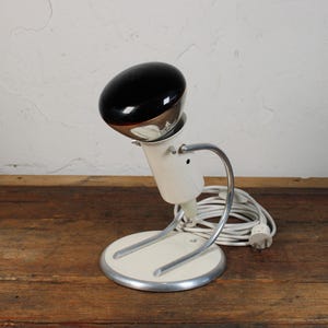 May include: Vintage white and chrome desk lamp with a black, oval-shaped head. The lamp has a circular base and a curved arm. A white power cord is coiled near the base. The lamp is on a wooden surface.