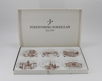 May include: A set of six white porcelain plates with brown illustrations of buildings, packaged in a white box. The box is labeled "FÜRSTENBERG PORZELLAN Seit 1747". Each plate features a different architectural design.