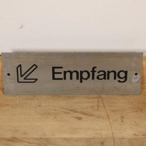 May include: A rectangular gray sign with the word "Empfang" in black lettering and a black arrow pointing left. The sign is set against a light-colored wall and a wooden surface.
