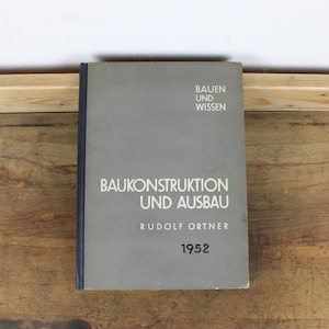 Peut inclure: Un livre vintage intitulé "BAUKONSTRUKTION UND AUSBAU" par Rudolf Ortner, publié en 1952. Le livre a une couverture grise avec du texte blanc et un dos bleu foncé. Le livre repose sur une surface en bois.