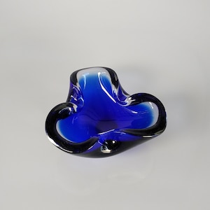 May include: A vibrant blue glass ashtray with a unique, organic shape. The ashtray features a deep, rich blue color with dark accents, creating a striking contrast. Its design includes curved edges and a smooth, glossy finish, perfect for home decor.