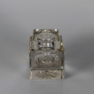 May include: A rectangular, clear glass inkwell with a silver-colored metal frame. The inkwell has a hinged lid and a decorative floral design on the front. The inkwell is likely vintage and suitable for calligraphy.