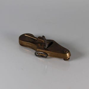 May include: A vintage brass shoe-shaped object, possibly a desk accessory or paperweight. The object has a detailed design with a small, oval-shaped ring on the side. The shoe is a warm, metallic color, and sits on a white surface.
