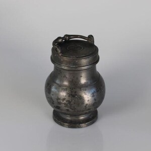 May include: An antique pewter jar with a lid and handle. The jar has a rounded body and a pedestal base. The lid is decorated with a small handle. The pewter has a dark, aged appearance.
