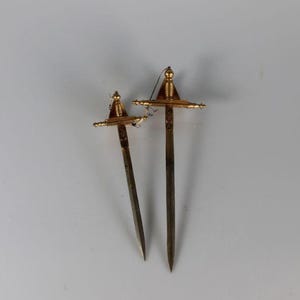 May include: Two decorative miniature swords with gold-colored hilts and dark blades are displayed against a white background. The swords are of different sizes, with intricate details on the hilt.