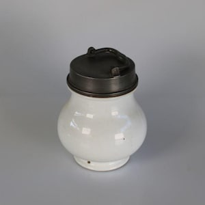 May include: A white ceramic jar with a dark metal lid. The jar has a rounded body and a small handle on the lid. The jar is likely used for storing food items.