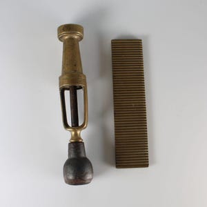 May include: An antique brass and wood wine bottle opener with a dark wooden handle and a separate grooved brass plate. The opener has a unique design with a cylindrical top and a cage-like structure.
