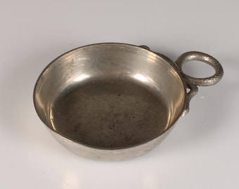 Peut inclure: Un petit bol rond en métal argenté avec une poignée. Le bol a un design simple et semble être fait d'une seule pièce de métal. La poignée est une petite boucle attachée au côté du bol. Le bol est posé sur une surface blanche.