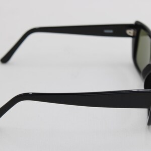 Puede incluir: Gafas de sol negras con montura rectangular y lentes oscuras. Las gafas tienen un dise&ntilde;o elegante y moderno con patillas negras gruesas y brillantes y un sutil detalle de bisagra. Las lentes parecen ser de color verde oscuro.