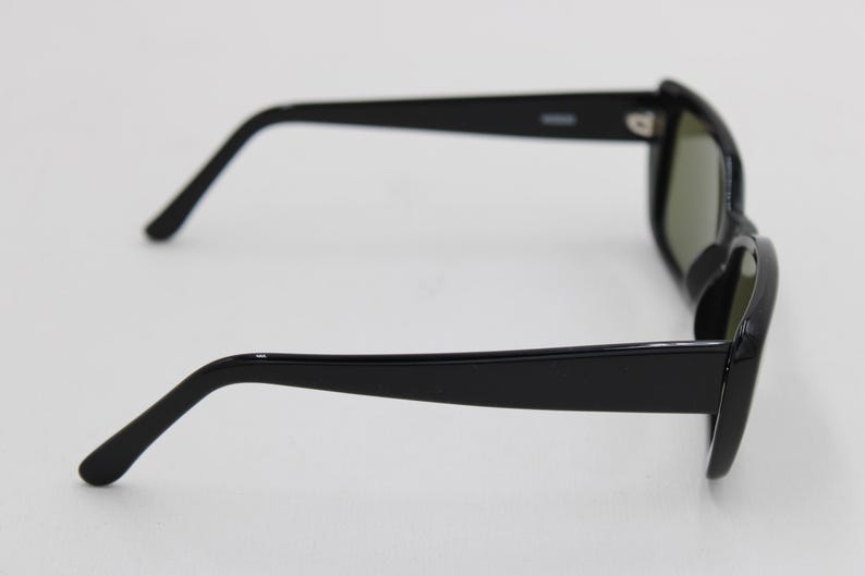 Puede incluir: Gafas de sol negras con montura rectangular y lentes oscuras. Las gafas tienen un dise&ntilde;o elegante y moderno con patillas negras gruesas y brillantes y un sutil detalle de bisagra. Las lentes parecen ser de color verde oscuro.