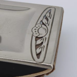 May include: Close-up of a silver-toned ink blotter with a decorative floral design. The blotter has a rounded handle and a rectangular base with a floral accent. The base is trimmed with a brown material.