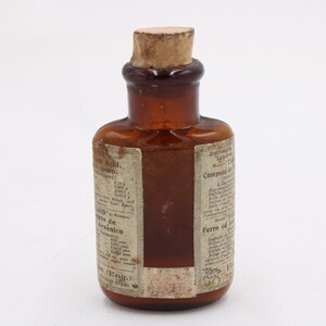 May include: A small, antique brown glass bottle with a cork stopper. The bottle has a label with faded text. The bottle is likely from the apothecary or medicine category.