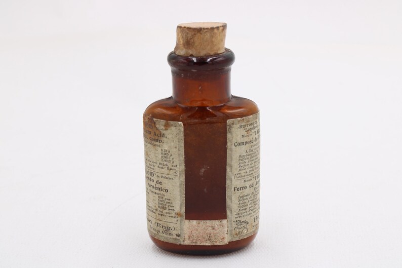May include: A small, antique brown glass bottle with a cork stopper. The bottle has a label with faded text. The bottle is likely from the apothecary or medicine category.