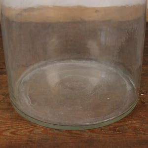 May include: A large, clear glass jar with a wide, cylindrical shape. The jar has a thick base and a slightly flared rim. The glass has a slightly cloudy appearance, and the jar sits on a wooden surface.