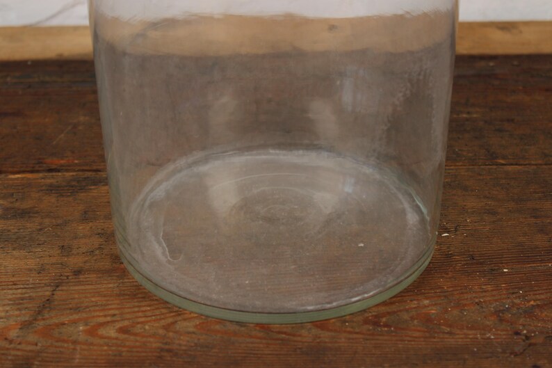 May include: A large, clear glass jar with a wide, cylindrical shape. The jar has a thick base and a slightly flared rim. The glass has a slightly cloudy appearance, and the jar sits on a wooden surface.