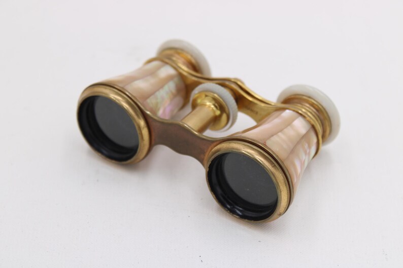 May include: Vintage opera glasses with a gold-tone frame and mother-of-pearl accents. The binoculars have black lenses and white end caps. A classic accessory for theater or events.