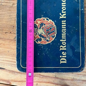 May include: A blue rectangular tin with the words "Die Rotmann Krone" in white. A pink and yellow measuring tape is placed on top of the tin. The tin has a decorative emblem in the center.