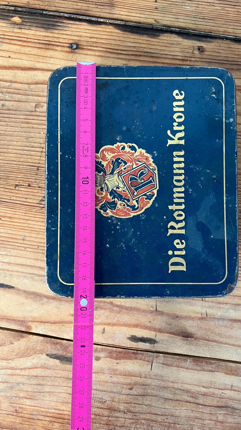May include: A blue rectangular tin with the words "Die Rotmann Krone" in white. A pink and yellow measuring tape is placed on top of the tin. The tin has a decorative emblem in the center.
