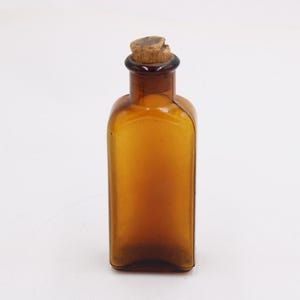 May include: Amber-colored glass bottle with a cork stopper. The bottle has a rectangular shape with rounded shoulders and a dark brown neck. The cork is a light brown color. The bottle is empty and sits on a white surface.