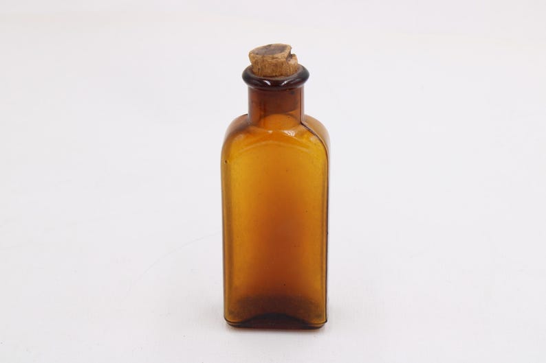 May include: Amber-colored glass bottle with a cork stopper. The bottle has a rectangular shape with rounded shoulders and a dark brown neck. The cork is a light brown color. The bottle is empty and sits on a white surface.