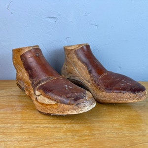 May include: Two brown leather shoe lasts, used for making shoes, are sitting on a wooden surface. The lasts are made of wood and leather and have a worn, vintage look.
