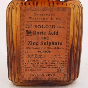 K&ouml;nnte beinhalten: Eine antike bernsteinfarbene Glasflasche mit einem Etikett mit der Aufschrift "SOLOID' BRAND Boric Acid and Zinc Sulphate". Das Etikett enth&auml;lt auch das Wort "POISON" und Gebrauchsanweisungen. Die Flasche stammt aus London, England.