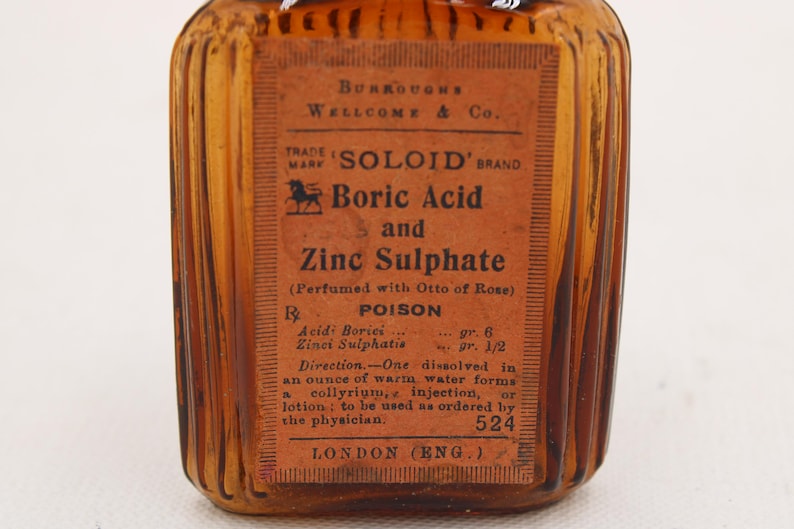 K&ouml;nnte beinhalten: Eine antike bernsteinfarbene Glasflasche mit einem Etikett mit der Aufschrift "SOLOID' BRAND Boric Acid and Zinc Sulphate". Das Etikett enth&auml;lt auch das Wort "POISON" und Gebrauchsanweisungen. Die Flasche stammt aus London, England.