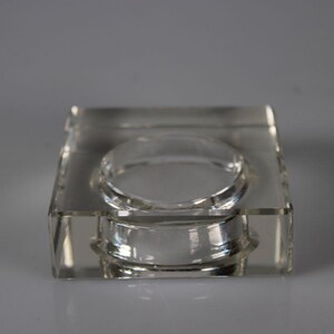 May include: A clear, square-shaped glass ashtray with a circular indentation in the center. The ashtray is made of thick, transparent glass and sits on a light gray background. The design is simple and modern.