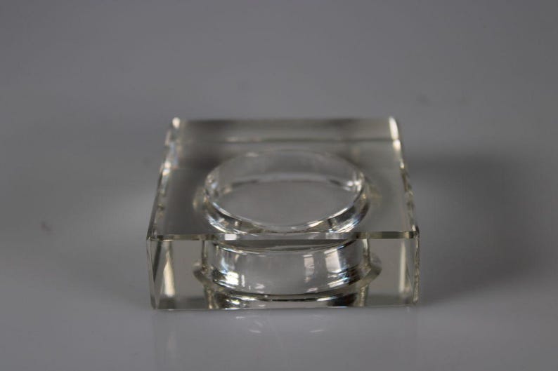 May include: A clear, square-shaped glass ashtray with a circular indentation in the center. The ashtray is made of thick, transparent glass and sits on a light gray background. The design is simple and modern.