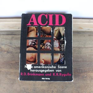 Può includere: Un libro intitolato "ACID" con copertina nera e scritte rosa. La copertina presenta una griglia di immagini. È visibile anche il testo "Die amerikanische Szene herausgegeben von R.D. Brinkmann und R.R. Rygulla".