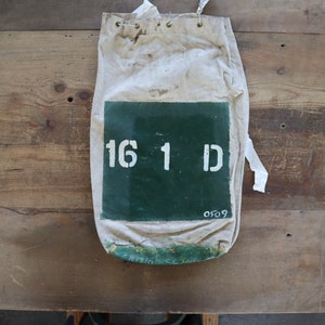 Puede incluir: Una bolsa de lona blanca con un parche verde que dice "16 1 D" y el número "0509" escrito debajo. La bolsa tiene un cierre de cordón y está hecha de un material resistente.